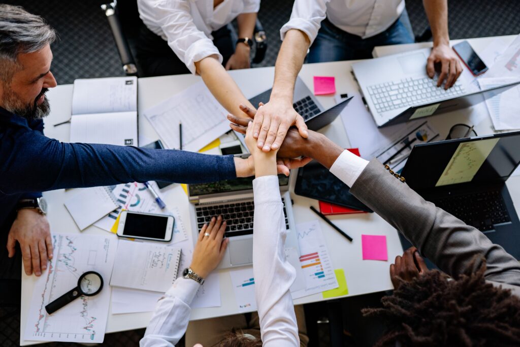 Group of people with their hands together over a table full of work. They appear to be celebrating team work.